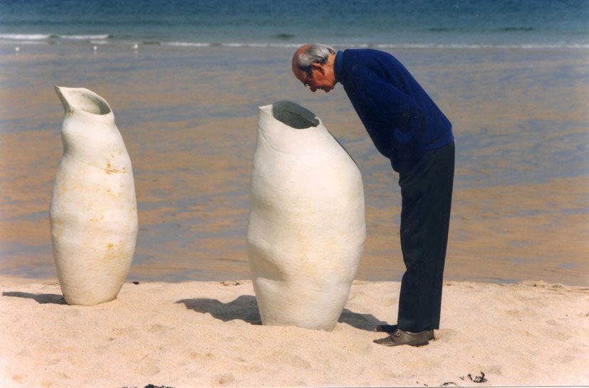 A man bending to look into a tall pale vessel on a sandy beach, with a second vessel beside it