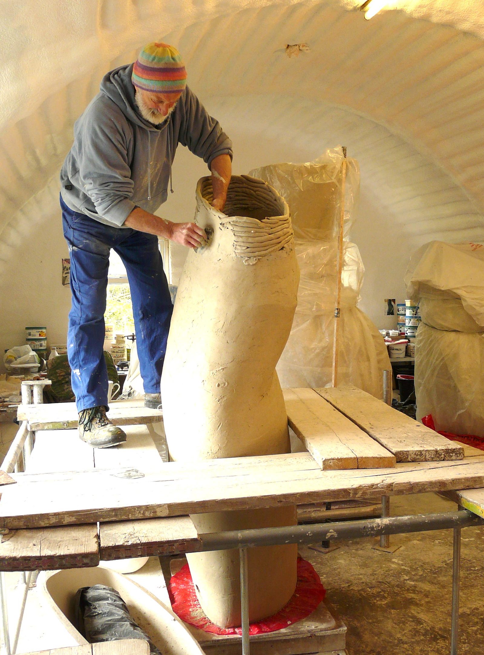 A man in a woolly hat smoothing the coiled rim of a large vessel in his studio