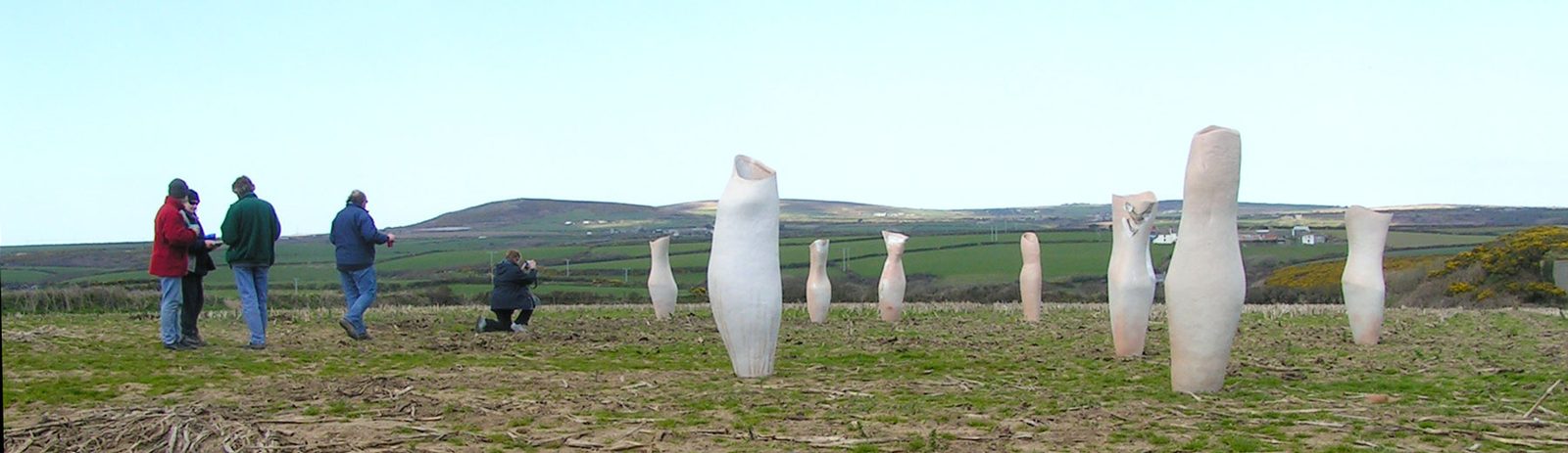 Tall pale vessels standing in a field of green hills, with a few visitors looking on
