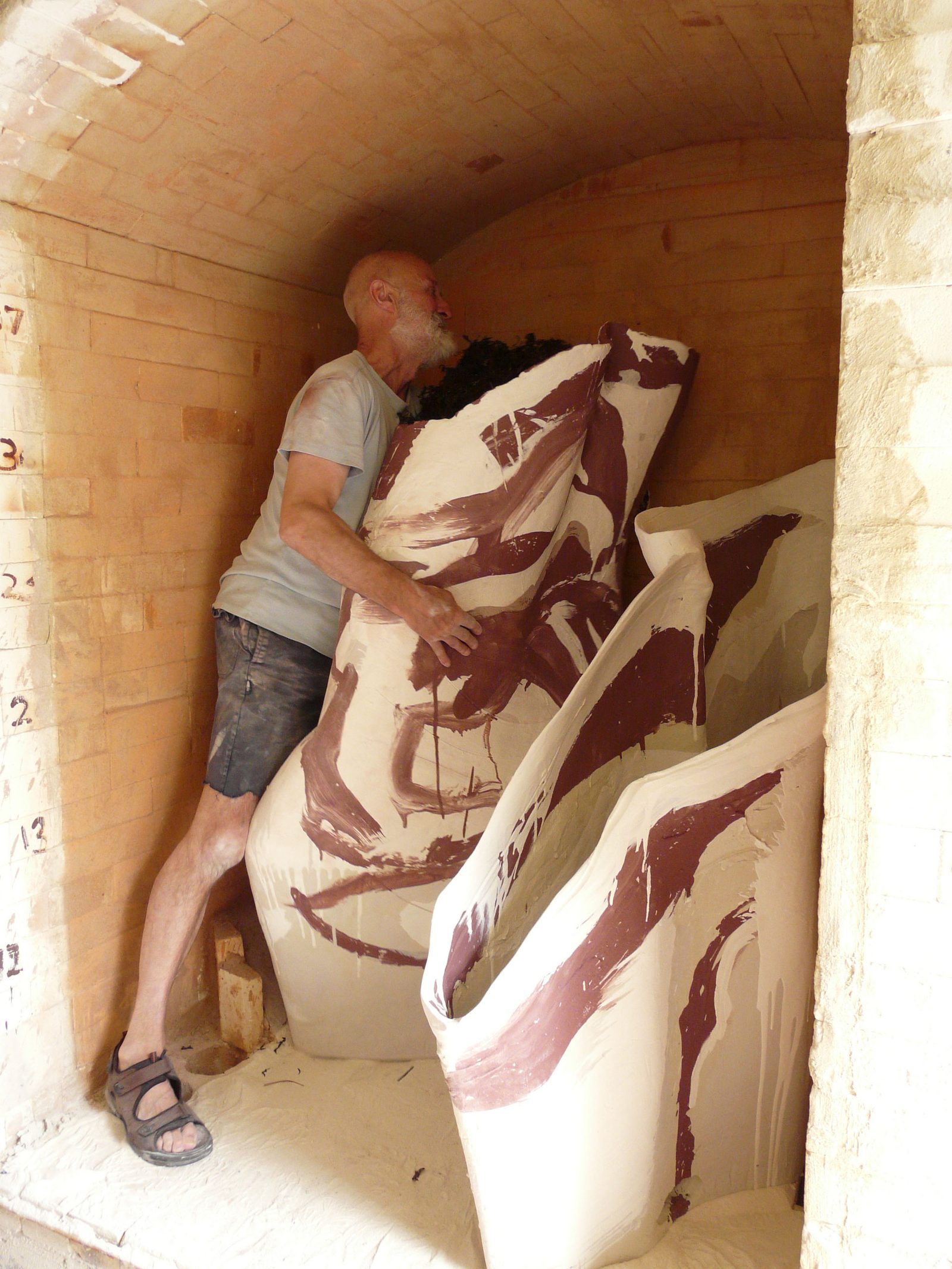 A man steadying a large vessel painted with maroon marks inside a brick kiln